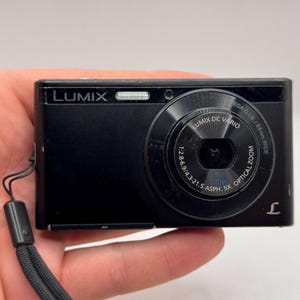 Puede incluir: Una cámara digital Lumix negra con un objetivo Lumix DC Vario que muestra las especificaciones 1:2.8-6.9/4.3-21.5 Asph. 5x optical zoom impresas en la parte frontal. El dispositivo compacto incluye una correa de muñeca negra y un logotipo.