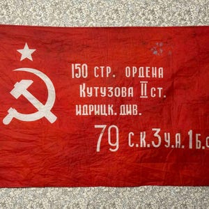Könnte beinhalten: Eine rechteckige rote Flagge mit einem weißen Hammer-und-Sichel-Symbol und einem Stern darüber auf der linken Seite. Der Stoff zeigt weißen kyrillischen Text mit der Aufschrift 150 СТР. ОРДЕНА КУТУЗОВА II СТ. ИДРИЦК. ДИВ. sowie 79 С.К. 3 У.А. 1 Б.Ф. in der Mitte.