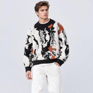 Op de afbeelding: Een trui met lange mouwen en een ronde hals met een abstract patroon in zwart, wit en gebrand oranje. Het gebreide kledingstuk heeft een effen zwarte geribbelde kraag, boorden en zoom, gecombineerd met een witte broek voor een hoog contrast.