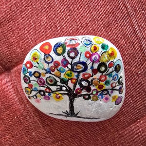 May include: A smooth, white, oval-shaped river rock featuring a hand-painted tree design. The tree has a black trunk and branches with numerous colourful, circular leaves in shades of red, yellow, blue, green, and gold, creating a vibrant pattern.