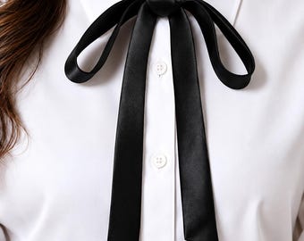 Puede incluir: Una camisa blanca de botones con cuello en punta y mangas largas. Una cinta de satén negro lisa está atada en un lazo en el cuello, con dos extremos largos y planos que cuelgan sobre la tapeta frontal de botones de la camisa.