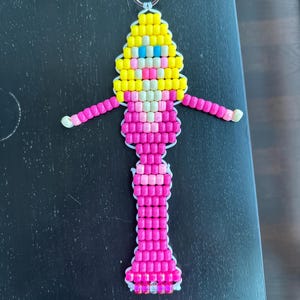 May include: A keychain crafted from plastic pony beads in the shape of a figure with yellow hair, blue eyes, and a pink outfit. The figure has outstretched arms and a long, vertical body design, all held together by white cord with a metal ring at the top.