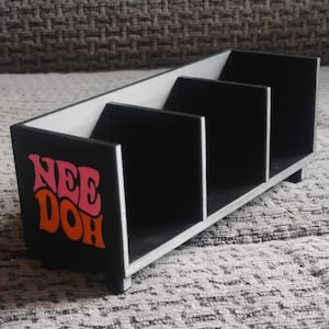 Op de afbeelding: Een rechthoekige zwarte opbergorganizer met drie interne compartimenten en vier kleine voetjes. Het voorpaneel is voorzien van de tekst "NEE DOH" in een gestileerd roze en oranje lettertype. De binnenwanden en de bodem zijn zwart met lichtgrijze randen.