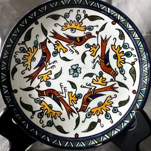 Könnte beinhalten: Ein runder Keramikteller mit weißem Hintergrund und einem handgemalten Muster aus vier orangefarbenen und blauen Vögeln, umgeben von gelben Blumenmotiven und grünen Blättern. Der Rand hat eine dunkelblaue Umrandung mit einem geometrischen grün-weißen Muster.