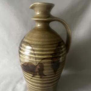 May include: A ceramic pitcher with a light brown, ribbed exterior and a glossy finish. The vessel features a narrow neck, a flared spout, and a single curved handle on the side. Dark brown, leaf-shaped patterns are painted around the body.