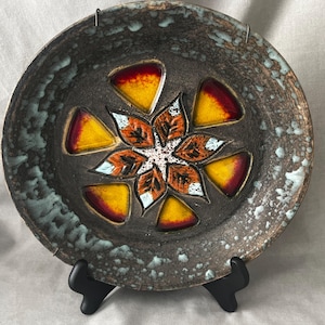 May include: A round ceramic plate with a dark brown, textured surface featuring a central floral motif of orange and white leaves surrounded by six triangular insets in gradient shades of yellow and red, with light blue glaze accents on the rim.