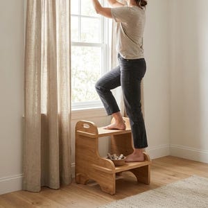 May include: A light-colored wooden two-step stool with a smooth finish and integrated handle cutouts on the top step. The stool features a sturdy, curved side design and is positioned on a wooden floor near a window with beige linen curtains.