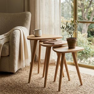 May include: A set of three nesting tables made of light-toned wood with rounded triangular tops and tapered legs. The tables are arranged in a stepped formation, with a ceramic mug on the tallest table and a small potted plant on the shortest.
