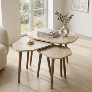 Könnte beinhalten: Ein Set aus drei Satztischen mit unregelmäßigen, abgerundeten dreieckigen Tischplatten in hellbeiger Marmoroptik und konischen Holzbeinen. Ein kleines goldenes Objekt in Apfelform steht auf dem kleinsten Tisch neben einer weißen Vase.