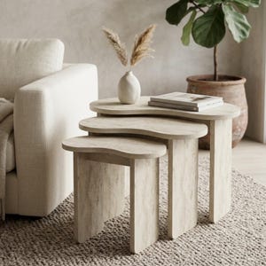May include: A set of three nesting tables with a light wood grain finish and organic, kidney-shaped tops. The tables feature curved, slab-style legs and are arranged in a cascading formation, with a white vase containing dried pampas grass on top.
