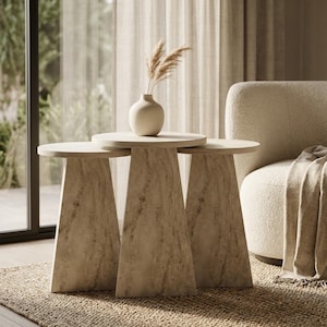 May include: A set of three nesting side tables with a beige marble-effect finish and tapered bases. The tables feature circular tops and a smooth, stone-textured surface. A small, round, off-white ceramic vase containing dried pampas grass sits on top.