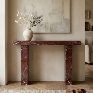 May include: A rectangular console table crafted from deep red marble with prominent white veining. The table features a flat top supported by two thick, vertical slab legs. A cream-coloured ceramic vase containing dried botanical stems sits on top.