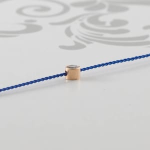 May include: A single, small, round, clear gemstone bead is strung on a blue cord. The bead is set in a gold-toned metal setting.