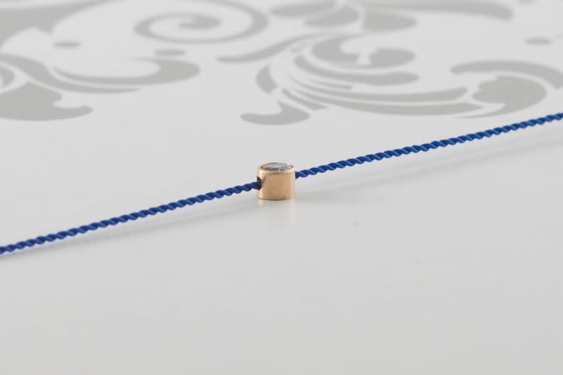 May include: A single, small, round, clear gemstone bead is strung on a blue cord. The bead is set in a gold-toned metal setting.