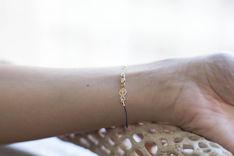 May include: A gold chain bracelet with a peace sign charm and a blue string tie closure.