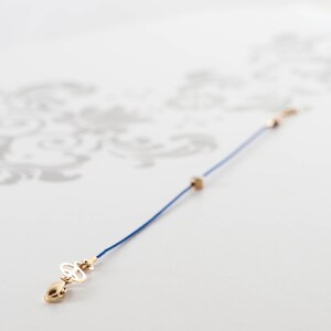May include: A blue cord bracelet with a gold charm featuring a symbol. The charm is attached to a small gold clasp.