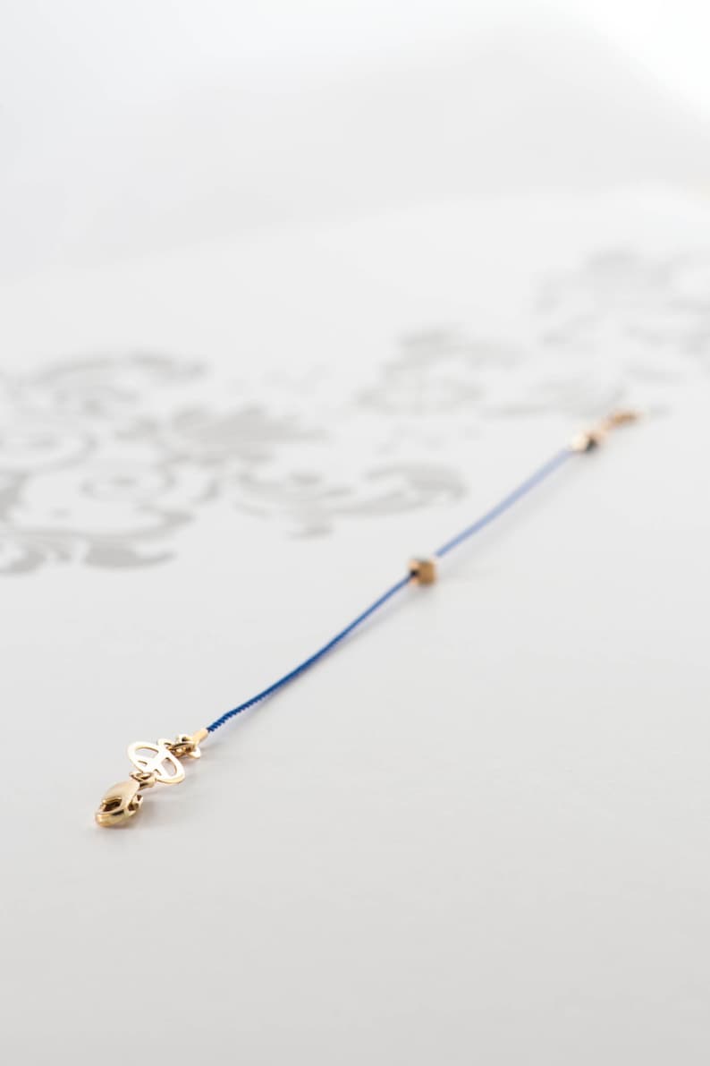 May include: A blue cord bracelet with a gold charm featuring a symbol. The charm is attached to a small gold clasp.
