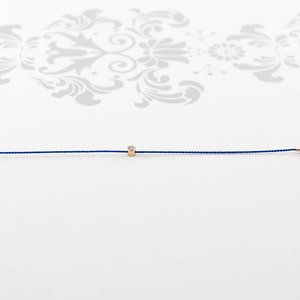 May include: A delicate blue string bracelet with a small gold bead featuring a tiny diamond.