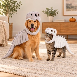 Könnte beinhalten: Ein Golden Retriever und eine getigerte Katze tragen passende hellgraue Plüsch-Dinosaurierkostüme. Jedes Kostüm besteht aus einer Kopfbedeckung mit weißen Zähnen, die das Gesicht umrahmen, und einem langen, segmentierten Schwanzteil.