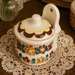 May include: A cream-colored ceramic wall-mounted salt crock with a matching lid. The container features hand-painted floral motifs in shades of brown, orange, and teal, with a scalloped yellow and brown pattern decorating the top of the lid.