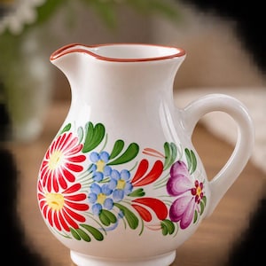Op de afbeelding: Een witte keramische kan met een handgeschilderd bloemmotief met rode, blauwe en paarse bloemen geaccentueerd door groene bladeren. De kan heeft een gebogen handvat, een brede tuit en een dunne rode rand langs de bovenkant van de opening.