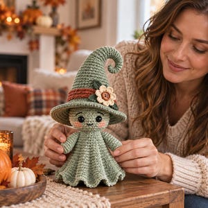 May include: A sage green crocheted doll wearing a tall, pointed witch hat with a curled tip. The hat features a rust-colored band and a small cream-colored flower with a brown button center. The doll has black bead eyes and pink cheeks.