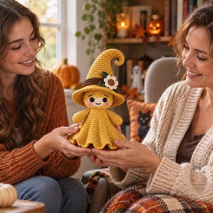 May include: A yellow crocheted doll wearing a pointed witch hat with a brown band and a white flower accent. The doll has a rounded head, small black eyes, pink cheeks, and a flared skirt base, all crafted with a textured yarn stitch pattern.