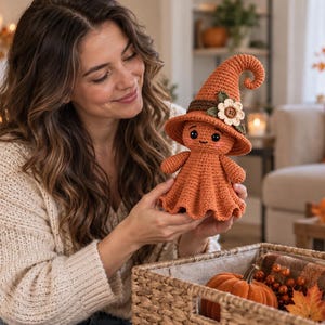 May include: A crocheted orange doll shaped like a ghost wearing a tall, pointed witch hat. The hat features a dark brown band, a small green leaf, and a cream-colored flower with a brown button center. The doll has black eyes and a small smile.
