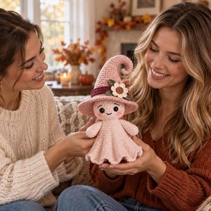 May include: A light pink crocheted ghost doll wearing a tall, pointed witch hat. The hat features a dark maroon band and a cream-colored flower with a brown center. The ghost has black embroidered eyes, a small smile, and rosy pink cheeks.