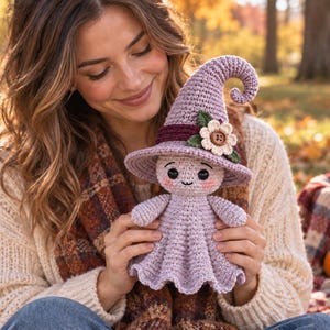 May include: A lavender crochet doll wearing a pointed witch hat with a dark purple band, a cream-colored flower with a brown button center, and green leaves. The doll has a round head, black eyes, a small stitched mouth, and pink cheeks.