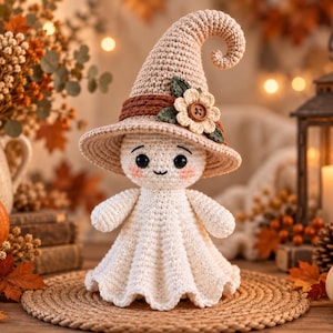 Puede incluir: Una muñeca fantasma de ganchillo blanca con un sombrero alto, puntiagudo y de color tostado, adornado con una banda marrón, una flor crema y hojas verdes. El fantasma tiene ojos de cuentas negras, boca pequeña y mejillas rosadas sobre una alfombra.