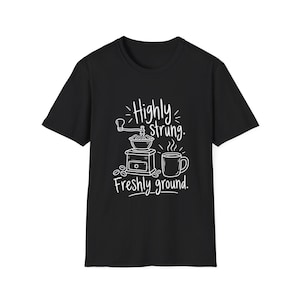 May include: A black short-sleeved t-shirt featuring a white graphic print of a manual coffee grinder, a steaming mug, and coffee beans. The text reads Highly strung. Freshly ground. The design is centered on the front of the garment.