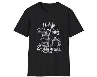 Könnte beinhalten: Ein schwarzes T-Shirt mit kurzen Ärmeln und einem weißen Grafikdruck, der eine manuelle Kaffeemühle, eine dampfende Tasse und Kaffeebohnen zeigt. Der Text lautet Highly strung. Freshly ground. Das Motiv ist mittig auf der Vorderseite platziert.