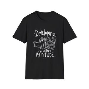 May include: A black short-sleeved t-shirt featuring a white graphic of a 35mm film canister and a strip of film negatives. The text "Developing a better ATTITUDE." is printed in white, stylized lettering above and below the film graphic.