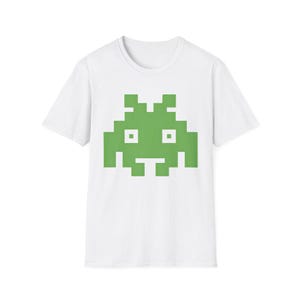 May include: A white short-sleeved t-shirt featuring a large, centered, pixelated alien graphic in a bright lime green color. The alien design is composed of square blocks and includes two white square eyes set within the green pixelated shape.