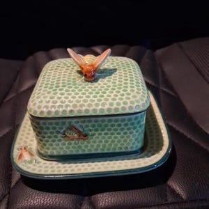 May include: A square ceramic butter dish with a lid featuring a light green honeycomb pattern. A three-dimensional bee figurine sits on top of the lid, and a small bee illustration is painted on the side of the base and the lid's edge.