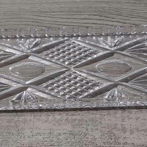 May include: A rectangular clear glass tray featuring a pressed pattern of diamond shapes, cross-hatched grids, and fan-like motifs. The edges of the tray are scalloped with a repeating notched design, creating a textured surface throughout the piece.