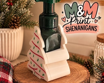 May include: A dark green plastic soap dispenser with a pump mechanism, held within a white, tree-shaped plastic holder featuring red and green decorative lines. The text M&M Print SHENANIGANS is displayed in the upper right corner in colorful font.