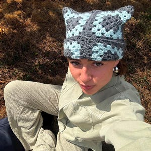 May include: A crochet beanie featuring a cat-ear silhouette, constructed from a series of square motifs in alternating shades of dark charcoal gray and light baby blue. The hat has a textured, chunky yarn appearance with a folded brim edge.