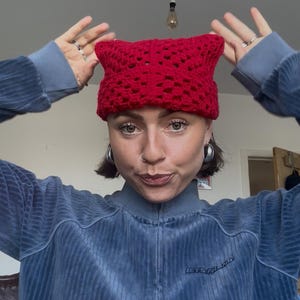 May include: A bright red crochet beanie featuring a distinct cat-ear silhouette. The hat is constructed with an openwork, grid-like stitch pattern throughout, creating a textured appearance. The design includes two pointed corners at the top.