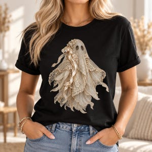 Peut inclure: Un t-shirt noir à manches courtes arborant un motif central représentant un fantôme drapé dans un tissu en dentelle couleur crème, orné de petites fleurs et de rubans délicats, créant un aspect superposé et texturé sur le tissu sombre.