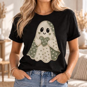 Può includere: Una t-shirt nera a maniche corte con una grafica centrale raffigurante un fantasma con un motivo a cuori color crema e verde salvia. Il fantasma ha grandi occhi neri, un piccolo sorriso e tiene un cuore verde salvia al centro.