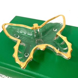 Op de afbeelding: Een heldere glazen vlindervormige schaal met een goudkleurige rand. De schaal ligt op een groen boek. De vlinderschaal heeft een delicaat ontwerp en wordt waarschijnlijk gebruikt voor decoratieve doeleinden.