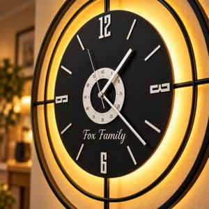 Könnte beinhalten: Eine runde schwarze Wanduhr mit Metallrahmen und warmer LED-Hintergrundbeleuchtung. Das Zifferblatt zeigt weiße Ziffern bei 12 und 6, silberne Zeiger, ein zentrales Zifferblatt mit Minutenmarkierungen und den weißen Schriftzug Fox Family.