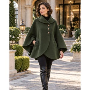 May include: An olive green wool-blend cape coat featuring a wide fold-over collar, three large round metallic buttons down the front, and two side pockets. The garment has a flared silhouette with open arm slits and a smooth, solid texture.