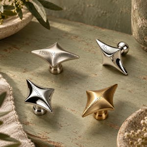 May include: Four star-shaped cabinet knobs with a pointed, geometric design. The set includes two polished silver-tone knobs, one brushed silver-tone knob, and one brushed gold-tone knob. Each knob features the text Abarrotesjuanki printed on the surface.