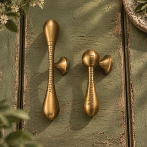 May include: Two brass-toned metal cabinet pulls with a smooth, elongated teardrop shape and a rounded base. Each handle features a central mounting point and a vertical orientation. The text Abarrotesjuanki is printed on the surface of both pulls.