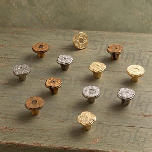 May include: A collection of eleven metal cabinet knobs in various finishes including antique brass, silver, and gold. Each knob features a circular or clover-shaped design with intricate embossed floral patterns on the top surface and a central post.