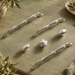 May include: A collection of polished silver-toned cabinet hardware including two long, wavy-shaped drawer pulls, two smooth oval-shaped knobs, and one small star-shaped knob, all featuring a reflective metallic finish and curved mounting points.