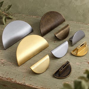 May include: A collection of semi-circular cabinet pulls in various sizes and finishes, including brushed gold, matte silver, and antique bronze. Each handle features a smooth, curved design with visible mounting points on the underside of some pieces.
